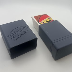 May include: A dark gray plastic storage case for a deck of UNO cards. The case consists of two rectangular pieces, one of which is empty and features the embossed UNO logo on its side, while the other holds a partial deck of UNO playing cards.