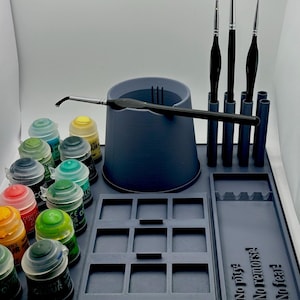 Può includere: Un organizer per hobby in plastica grigio scuro con un portabicchieri centrale, una griglia a nove scomparti e un vassoio rettangolare con il testo in rilievo No pity! No remorse! No fear! Include tre pennelli in posizione verticale.