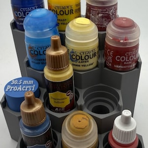 Könnte beinhalten: Ein graues, abgestuftes hexagonales Farbregal mit verschiedenen Hobby-Farbflaschen. Die Sammlung umfasst Citadel Colour-Töpfe in Blau, Gelb und Rot sowie Tropfflaschen mit den Aufschriften ProAcryl, Army Painter Daemonic Yellow, Ultramarine Blue und Dragonfire Red.