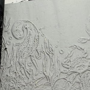 Könnte beinhalten: Eine hellgraue, strukturierte Reliefplatte mit einer geprägten Unterwasserszene, die ein Seepferdchen, eine Krabbe, Seetang und Blasen zeigt. Links zeigt ein schwarzer Druckstock dasselbe komplexe Meeresmotiv im kontrastreichen Negativformat.