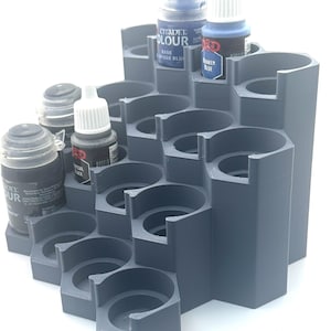 Può includere: Un organizer per boccette di vernice grigio, a gradini, stampato in 3D con una struttura ispirata agli alveari. Presenta molteplici alloggiamenti circolari per piccoli barattoli di vernice, inclusi flaconi Citadel Colour etichettati Base Macragge Blue e Abaddon Black.