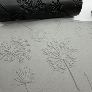 May include: A black cylindrical rolling pin with a carved dandelion pattern positioned above a flat, light grey slab of clay. The rolling pin features recessed floral designs that create a matching embossed impression on the surface of the clay.