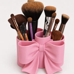 Peut inclure: Un porte-pinceaux de maquillage cylindrique rose clair avec un nœud moulé proéminent sur le devant. Le récipient comporte plusieurs compartiments intérieurs remplis de divers pinceaux de maquillage de tailles, formes et couleurs variées.