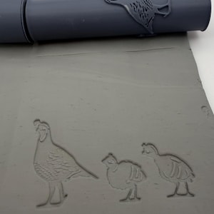 May include: A grey cylindrical rolling pin with a raised quail design, positioned above a flat sheet of grey clay that features an impressed pattern of one adult quail and two smaller quail chicks, creating a clear textured relief in the clay surface.