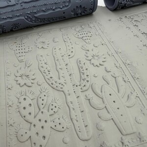 May include: A gray cylindrical clay texture roller featuring a raised pattern of various cacti, desert flowers, and small stars. The roller is positioned above a flat slab of light gray clay that displays the same embossed desert-themed design.