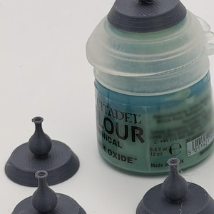 May include: Three small, dark gray, 3D-printed vase-shaped objects with circular bases and narrow necks, arranged near a translucent plastic Citadel Colour paint pot labeled Nihilakh Oxide, which contains 0.4 fl oz of paint.
