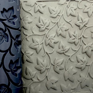May include: A dark gray cylindrical rolling pin with a recessed ivy leaf pattern positioned next to a flat slab of light gray clay that has been impressed with the same repeating ivy leaf design, showing the texture created by the tool.