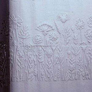 Pode incluir: Um rolo cilíndrico escuro com um padrão floral gravado é colocado ao lado de uma superfície de argila clara que exibe o mesmo design de flores silvestres em relevo, com vários caules, folhas e cabeças de flores circulares.