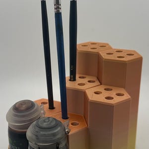 May include: A 3D-printed hexagonal paint brush and paint pot organiser featuring a gradient colour scheme transitioning from peach at the top to yellow at the base. The stand holds three paint brushes and two Citadel paint pots with clear lids.