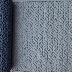 May include: A dark grey cylindrical rolling pin with a repeating embossed pattern of teardrop-shaped motifs. The roller is positioned next to a flat, light grey surface that displays the same intricate, pressed teardrop design in relief.