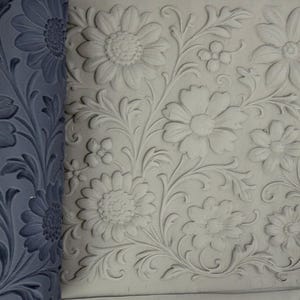 Può includere: Un mattarello in silicone grigio scuro con un motivo floreale profondo di margherite e viticci, accanto a una lastra di argilla bianca impressa con lo stesso intricato e ripetitivo disegno botanico di fiori e foglie.