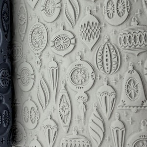 May include: A dark gray cylindrical rolling pin with a recessed pattern of various Christmas ornaments and small stars, shown next to a light gray slab of clay that has been impressed with the same detailed holiday bauble and star design.