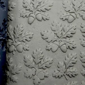 May include: A dark grey silicone texture mat featuring a repeating embossed pattern of oak leaves and acorns. The mat is shown pressed into a light grey clay surface, creating a raised, detailed impression of the botanical design.