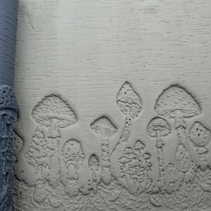 May include: A gray cylindrical rolling pin with a raised mushroom pattern and a flat slab of light gray clay showing the same embossed mushroom design. The texture features various mushroom shapes, including tall and short caps with gills.
