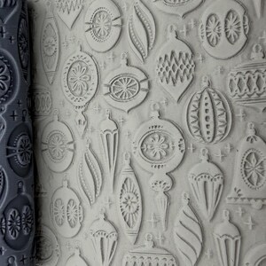 May include: A grey cylindrical rolling pin with a deeply engraved pattern of various Christmas ornaments and small stars, shown next to a flat piece of light-coloured clay that has been embossed with the same repeating holiday design.