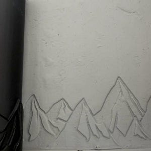 Può includere: Un mattarello cilindrico nero per argilla con un motivo a catena montuosa in rilievo, posizionato accanto a una lastra di argilla grigio chiaro che mostra lo stesso design montuoso impresso sulla superficie come un paesaggio continuo.