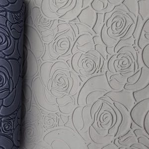Könnte beinhalten: Eine dunkelgraue zylindrische Teigrolle mit einem tiefen, sich wiederholenden Rosenmuster, neben einer hellgrauen Tonoberfläche, die dasselbe florale Design in das Material eingeprägt zeigt und eine klare, durchgehende Textur erzeugt.