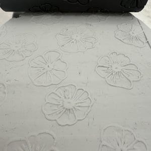 Op de afbeelding: Een zwarte cilindrische deegroller met een verdiept bloemmotief drukt in een platte, lichtgrijze kleiplaat. De deegroller laat een duidelijke, reliëfafdruk achter van meerdere vijfbladige bloemen op het oppervlak van de klei.