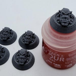 May include: Four dark grey, circular miniature bases featuring sculpted orc heads with prominent tusks and detailed facial features, alongside a translucent plastic paint pot labelled Citadel Colour with red paint and a volume of 12 ml.