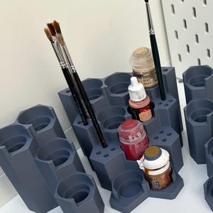 May include: A dark grey modular paint rack with a hexagonal grid design featuring multiple circular slots for paint pots and small holes for brushes. Several paint pots in various colours and thin paint brushes are inserted into the organiser.