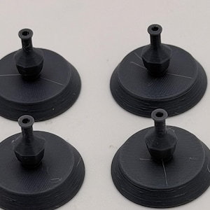 May include: Four black 3D printed objects, each consisting of a circular base with a small, narrow-necked vase-like structure centered on top. The surfaces of these items show fine horizontal layer lines characteristic of the additive manufacturing process.