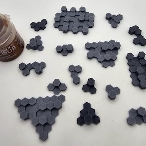 May include: A collection of dark gray 3D printed hexagonal terrain tiles in various cluster sizes and a brown Citadel Colour paint pot. The tiles feature a flat top surface and are arranged in multiple groupings across the frame.