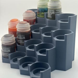 May include: A grey, tiered, 3D-printed paint rack with multiple circular slots designed to hold small paint pots. Several Citadel Colour paint pots in various colours are inserted into the upper levels of the organiser, which features a stepped, modular design for storage.
