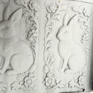 May include: A white silicone rolling pin featuring a repeating embossed pattern of two rabbits sitting upright, surrounded by intricate floral and leafy vine motifs. The design is carved into the surface to create a textured impression.