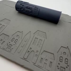 May include: A dark gray cylindrical rolling pin with embossed architectural details, positioned above a flat gray slab of clay that features a repeating, pressed pattern of various house designs including gabled roofs, windows, and arched doorways.