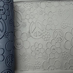 Op de afbeelding: Een donkergrijze cilindrische kleitextuurroller met een herhalend reliëfpatroon van lachende bloemen, vredestekens en kleine bloesems, die het ontwerp in een platte, lichtgrijze plaat van kneedbare klei drukt.
