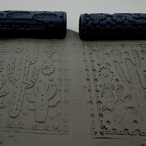 Peut inclure: Deux rouleaux cylindriques bleu foncé à motifs placés au-dessus de deux plaques d'argile grise rectangulaires. Chaque rouleau présente des motifs en relief de cactus et de fleurs, imprimés dans l'argile pour créer des textures.