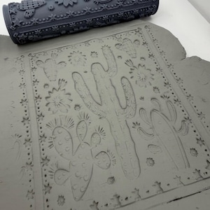 May include: A dark gray cylindrical clay rolling pin featuring a raised pattern of cacti, flowers, and stars. The roller is positioned above a flat slab of gray clay that has been impressed with the same desert-themed design, showing clear indentations.