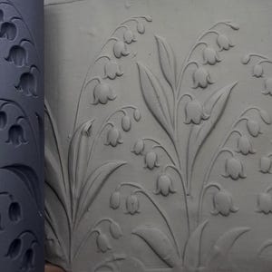 Könnte beinhalten: Eine dunkelgraue, zylindrische Ton-Texturwalze mit einem vertieften Maiglöckchen-Muster, platziert neben einer flachen Platte aus hellgrauem Ton, die dasselbe florale Design mit klaren, erhabenen Details auf ihrer Oberfläche eingeprägt zeigt.