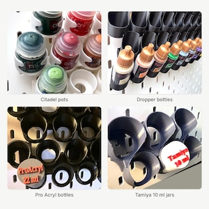 May include: A collection of white and black plastic wall-mounted organizers designed for hobby paint containers. The set includes holders for Citadel pots, dropper bottles, Pro Acryl 22 ml bottles, and Tamiya 10 ml jars, all mounted on a pegboard system.