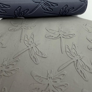 May include: A dark grey cylindrical rolling pin featuring a recessed dragonfly pattern. The tool is positioned above a flat, light grey slab of clay that displays a clear, embossed impression of the same dragonfly design across its surface.