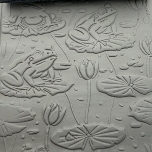 May include: A grey textured rolling pin with a carved pattern of frogs sitting on lily pads and lotus flowers, shown pressing into a flat slab of light grey clay to create a repeating embossed design of the same pond scene.