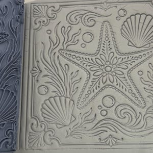 May include: A grey silicone rolling pin with a raised pattern of a starfish, seashells, and seaweed, shown next to a light grey clay slab that has been imprinted with the same detailed nautical design featuring bubbles and decorative scrollwork.