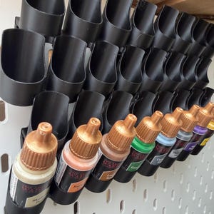 Op de afbeelding: Een zwart kunststof wandrek voor verf met meerdere cilindrische sleuven. Verschillende kleine plastic druppelflesjes met goudkleurige doppen en etiketten van The Army Painter zijn in de onderste rij van het rek geplaatst met diverse verfkleuren.