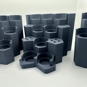 Op de afbeelding: Een donkergrijze, 3D-geprinte modulaire opbergorganizer met een gelaagd, zeshoekig ontwerp. De unit bevat meerdere cilindrische compartimenten van verschillende hoogtes en kleinere ronde gaten voor het rechtop bewaren van gereedschap of accessoires.