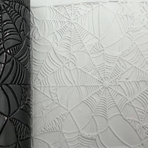 May include: A black cylindrical rolling pin featuring a recessed spider web pattern, positioned next to a white clay surface that displays the same spider web design embossed into the material, showing the texture and depth of the pattern.