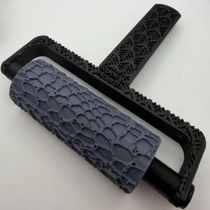 May include: A black plastic handle with an intricate lattice pattern and the text Rook & Hearth, attached to a grey cylindrical roller featuring a raised, organic, web-like texture designed for creating impressions on soft surfaces.