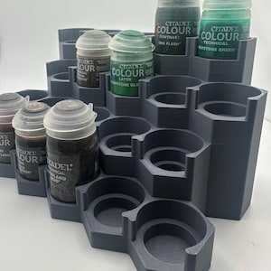 May include: A dark gray tiered paint rack with a hexagonal grid design, featuring multiple circular cutouts for holding paint pots. Several Citadel Colour paint pots with various labels are inserted into the slots of the plastic organizer.