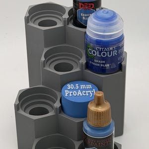 Könnte beinhalten: Ein grauer, abgestufter, sechseckiger Farbflaschen-Organizer mit mehreren runden Steckplätzen. Drei Farbflaschen sind eingesetzt, darunter D&D Kraken Blue, Citadel Colour Tyran Blue und Army Painter Ultramarine Blue. Ein blauer Zylinder mit der Aufschrift 30.5 mm ProAcryl ist ebenfalls vorhanden.