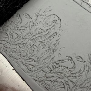 Op de afbeelding: Een lichtgrijze kleiplaat met een reliëf van een onderwaterscène. Het ontwerp bevat een grote inktvistentakel, een krab, zeewier en kleine belletjes. Het oppervlak heeft een matte textuur met diepe inkepingen die de afbeelding vormen.