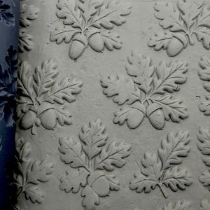 May include: A dark blue silicone mold featuring a repeating pattern of oak leaves and acorns, shown alongside a light gray clay impression that displays the raised, detailed relief of the botanical design in a consistent, tiled arrangement.