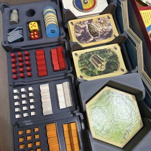 May include: A board game organiser tray containing various game components including red, white, and orange wooden cubes, two dice, a stack of blue circular tokens, and several decks of illustrated cards featuring landscapes and a circular shield design.