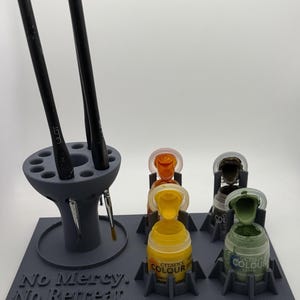 May include: A grey 3D-printed paint and brush organiser stand featuring a circular brush holder with multiple slots and a rectangular base with four paint pot holders. The base has the text "No Mercy. No Retreat." embossed on the front.