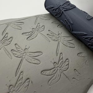 Könnte beinhalten: Eine dunkelgraue zylindrische Tonwalze mit erhabenem Libellenmuster, platziert neben einer hellgrauen Tonplatte, die mit demselben Libellendesign geprägt wurde, wodurch ein sich wiederholendes Insektenmuster auf der Oberfläche entsteht.