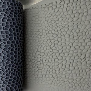 Könnte beinhalten: Eine dunkelgraue, zylindrische Strukturwalze mit einem erhabenen, unregelmäßigen, geometrischen Wabenmuster. Daneben liegt eine flache, hellgraue Tonplatte, die mit demselben sich wiederholenden, organischen, mehrseitigen Zellmuster geprägt ist.