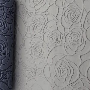 Könnte beinhalten: Eine dunkelgraue zylindrische Teigrolle mit tief eingeprägtem Rosenmuster, platziert neben einer hellgrauen Tonplatte, die mit demselben Rosendesign bedruckt wurde und die Texturübertragung von der Rolle auf die Oberfläche zeigt.